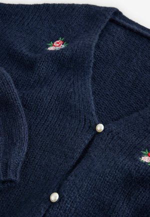 Navy zilā adīta kardigāna jaciņa ar V-veida kaklu, pērļu pogām un ziedu izšuvumiem ar rozā un zaļām akcentiem. Mīksta tekstūra un vidēja biezuma.