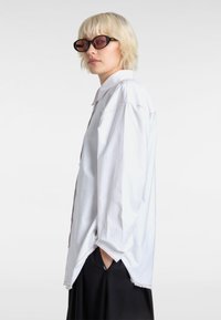White oversized button-up shirt with pink stitching, side slits, and long sleeves. Model wears black sunglasses and black bottom.