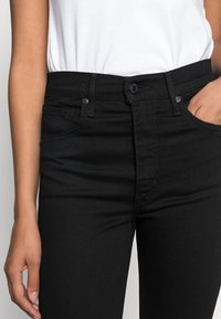 Mujer con jeans negros de cintura alta y una blusa blanca metida, con una mano parcialmente en el bolsillo delantero.