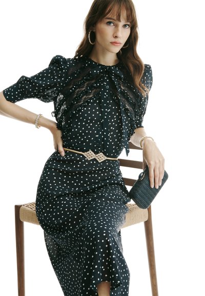 Ženska sedi na leseni stolici, oblečena v črno obleko s pikami in čipkastimi detajli, zlati pas, obroči za ušesa, zapestnice in drži teksturirano torbico.