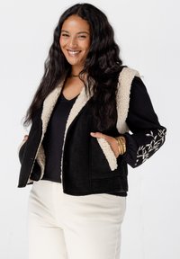 Zwarte vest met een crèmekleurige nepbont kraag en voering. Heeft zijzakken en een zwart geborduurd bloemenontwerp op de rechtermouw.