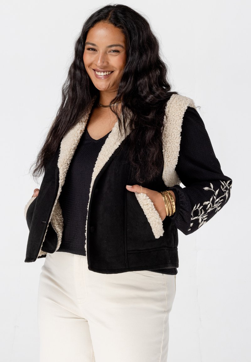 Zwarte vest met een crèmekleurige nepbont kraag en voering. Heeft zijzakken en een zwart geborduurd bloemenontwerp op de rechtermouw.