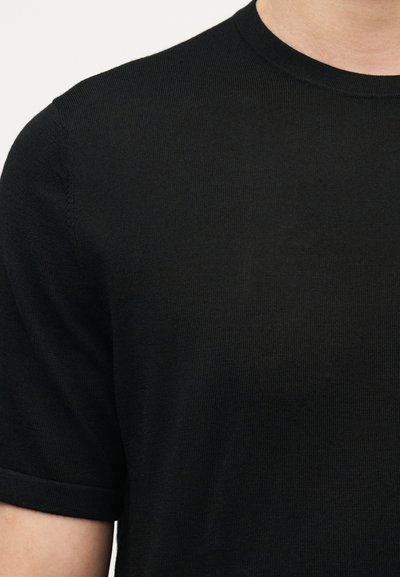 Κοντινό πλάνο ενός ατόμου που φοράει μονόχρωμο μαύρο κοντομάνικο πουλόβερ με στρογγυλή λαιμόκοψη και ομαλή, πλεκτή υφή.