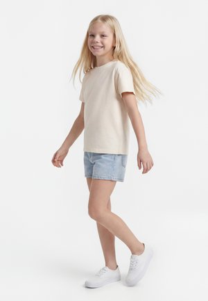 Ragazza bionda sorridente che cammina, indossa una maglietta beige, pantaloncini di jeans azzurri e scarpe da ginnastica bianche, su sfondo bianco.