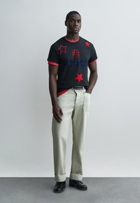 Hombre de pie con las manos en los bolsillos, vistiendo una camiseta negra con estrellas rojas y pantalones beige, sobre un fondo liso de color gris claro.