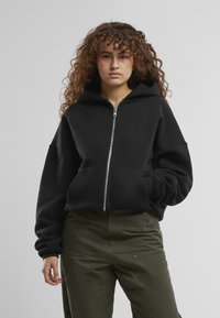 Schwarze Zip-Hoodie aus weichem Stoff, mit einer großen Kapuze, überschnittenen Schultern und gerippten Bündchen. Kombiniert mit olivgrünen Hosen.