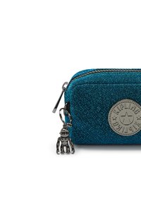 Türkis-glitzernde Tasche mit einem runden, eingeprägten Logo, silberner Hardware und einem Affen-Schlüsselanhänger. Reißverschlussverschluss oben. Kompaktes Design.