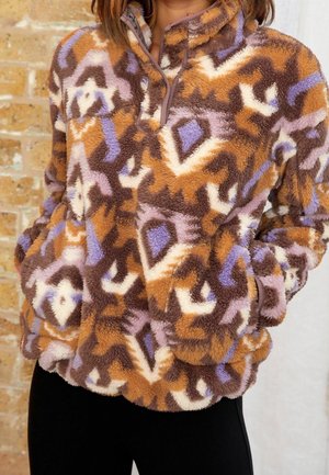 Eine Frau trägt einen braunen, lila und cremefarben gemusterten flauschigen Pullover mit einem Reißverschluss vorne und hat die Hände in den Taschen. Sie steht vor einer Ziegelwand.