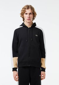 Čierna mikina s kapucňou na zips s bielymi a béžovými rukávmi, so predným klokaním vreckom a malým zeleným logom na hrudi. Mäkko tkaný materiál.