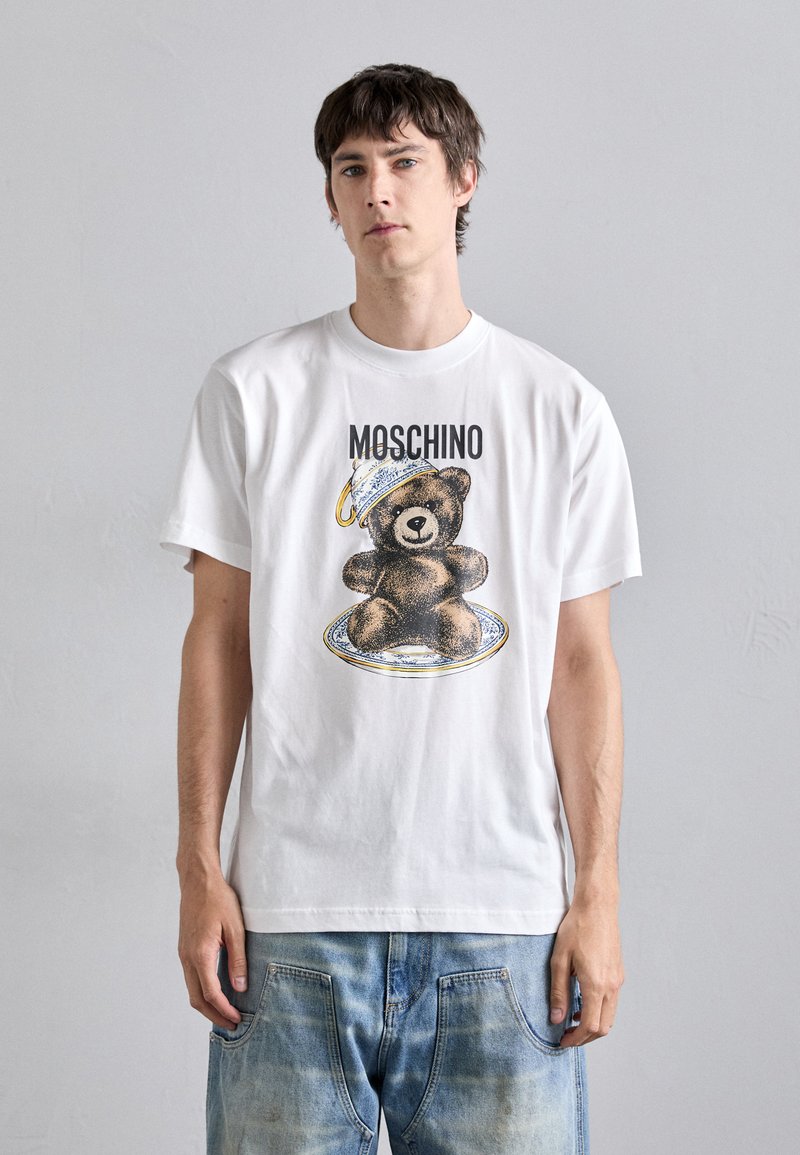 Balta marškinėliai su lokio grafika ant lėkštės, viršuje esantis tekstas "Moschino". Dizainas apima išsamią tekstūrą ir šviesias spalvas.