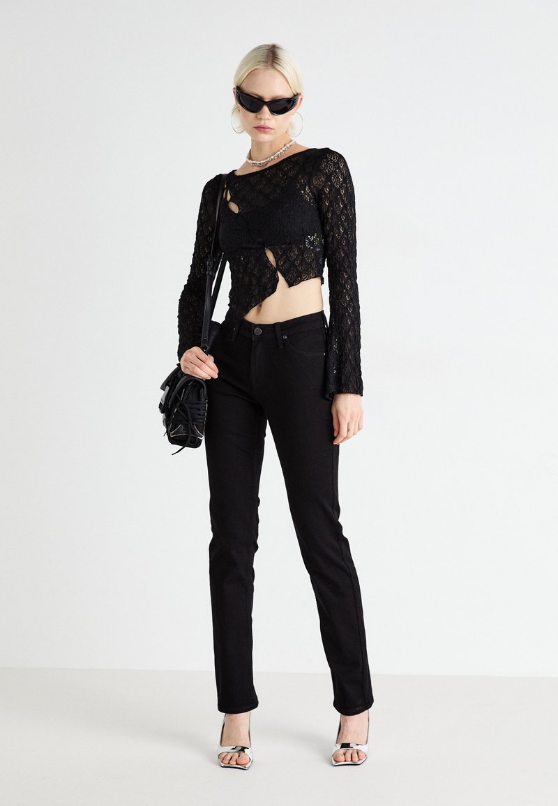 Haut en dentelle noire à manches longues avec découpes, associé à un jean noir ajusté. Les accessoires comprennent un petit sac noir et des sandales à talons transparents.
