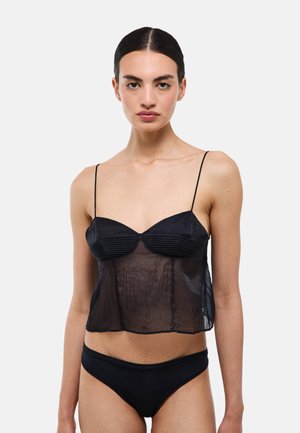 Femme aux cheveux foncés attachés porte un haut noir transparent à fines bretelles et un sous-vêtement noir, debout devant un fond clair uni.