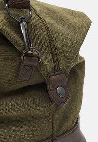 Mochila de lona verde oliva con solapa, que presenta un acento de cuero marrón, herrajes metálicos y un cierre de cremallera en la parte superior.