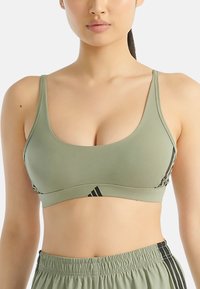 Olive zelená sportovní podprsenka s tenkými ramínky, výstřihem ve tvaru lodi a černými akcenty. Měkká tkanina má minimální švy a hladký povrch.