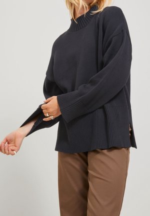 Femme ajustant la manche d'un pull en maille côtelée bleu marine foncé, portant un pantalon marron, avec une vue partielle des cheveux blonds et un fond neutre.