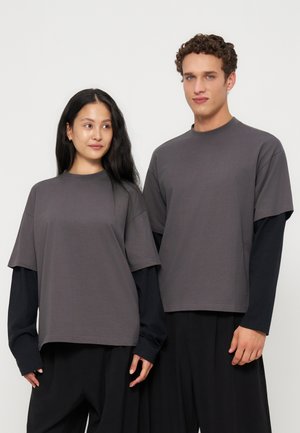 To modeller iført lagdelte sorte langærmede under løse grå kortærmede skjorter og sorte brede bukser, stående mod en hvid baggrund.