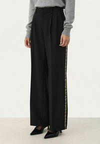 Zwarte wijde broek met plooien, voorzien van een gouden gestreepte zijkant. Gemaakt van een soepele stof, gecombineerd met zwarte puntige hakken.