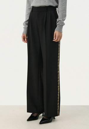Zwarte wijde broek met plooien, voorzien van een gouden gestreepte zijkant. Gemaakt van een soepele stof, gecombineerd met zwarte puntige hakken.