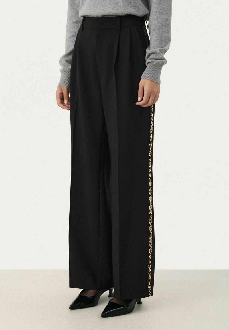 Zwarte wijde broek met plooien, voorzien van een gouden gestreepte zijkant. Gemaakt van een soepele stof, gecombineerd met zwarte puntige hakken.