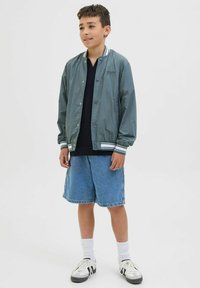 Grå lättviktsjacka med vitrandiga manschetter över en mörk skjorta, kombinerad med denimshorts och vita strumpor, kompletterat med svart- och gråsnickers.