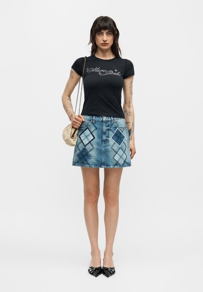 Γυναίκα με σκούρα μαλλιά μήκους μέχρι τους ώμους που φοράει μαύρο εφαρμοστό μπλουζάκι, τζιν φούστα με διαμαντένια σχέδια, μαύρα μυτερά παπούτσια και κρατάει μια πλεκτή τσάντα.