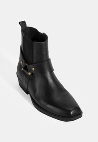 Bottines en cuir noir avec un bout pointu, texture lisse, accents dorés et une fermeture éclair sur le côté. Présente une sangle décorative avec des éléments en métal.