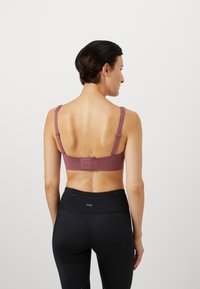 Kvinne med kort mørkt hår iført en mauve BH med justerbare stropper og svarte høyt liv leggings, vist fra bakgrunnen mot en hvit bakgrunn.