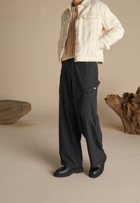 Chaqueta vaquera color crema claro, suéter de punto color tostado, pantalones anchos negros con bolsillos y zapatos negros gruesos, todo junto a un fondo neutro.