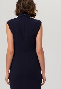 Marinblå klänning med korta ärmar, mjuk textur och synlig dragkedja på ryggen. Skräddarsydd silhuett med sömdetaljer längs bysten.