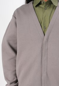 Šedý kardigán s hlubokým výstřihem do V, vyrobený z měkkého materiálu. Nosený přes olivově zelenou košili s knoflíkovými detaily a límcem.