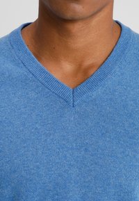 Nacke och bröst av en person med medelhudton som bär en ljusblå V-ringad stickad tröja mot en neutral bakgrund.