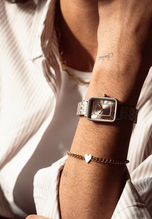Montre carrée argentée avec un bracelet métallique, présentant un cadran de couleur claire et des chiffres romains. Un bracelet en chaîne dorée avec un charme en forme de cœur est également visible.