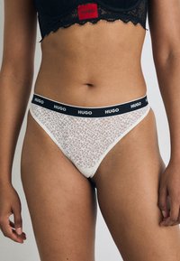 White lace thong with a floral pattern, featuring a black elastic waistband labeled "HUGO". Slim design, soft texture, and minimal coverage.