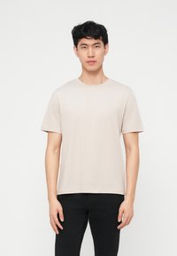Jeune homme aux cheveux noirs courts, portant un t-shirt beige clair uni à manches courtes et un pantalon noir, debout devant un fond blanc.