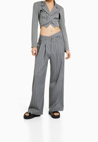 Femme portant un blazer cropped à rayures grises avec un détail découpé et un pantalon large assorti, associé à des sandales à plateforme noires.