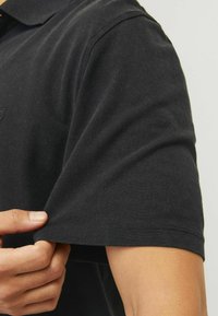 Čierna polo košeľa s textúrovanou látkou. Detail na rukáve s rukou ťahajúcou manžetu. Krátke rukávy a klasický golier.