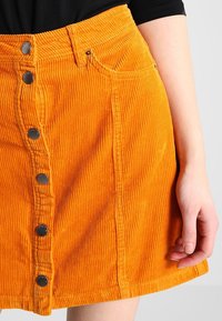 Jupe en velours côtelé orange avec nervures verticales, fermeture avant à boutons, deux poches latérales et coupe droite.