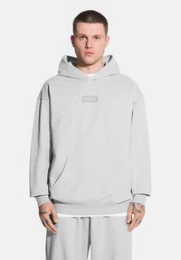 Mężczyzna stoi na białym tle, ubrany w szary sweatshirt z logo oraz szare dresy. Ma krótkie włosy i tatuaże na rękach.