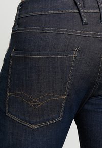 Tmavě modré džínové kalhoty s zadní kapsou zdobenou výrazným cikcak stehem a kontrastními hnědými nitěmi.