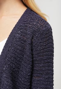 Cárdigan de punto en azul marino oscuro. Presenta un frente abierto, un corte holgado y un patrón texturizado con anchos variables y una ligera transparencia.