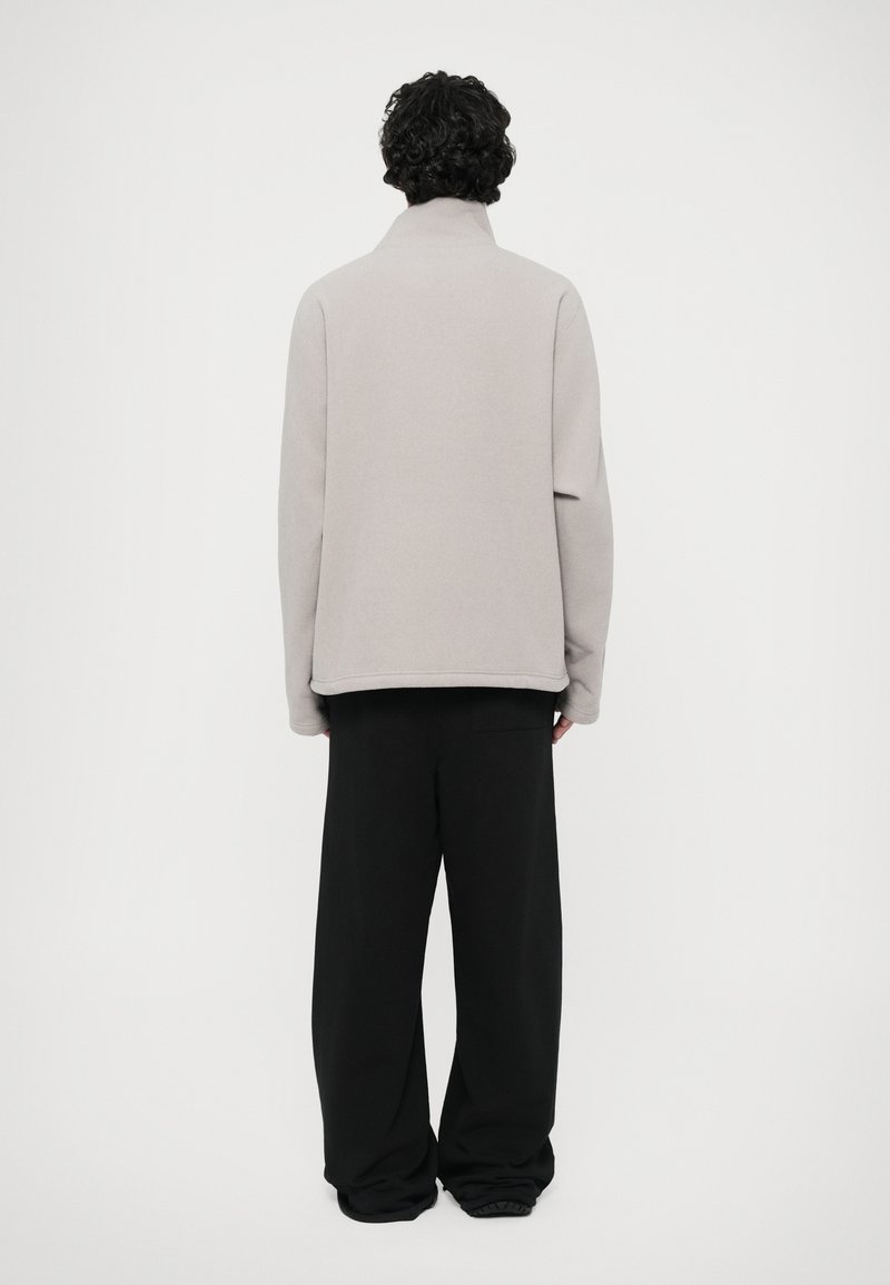 Light gray sweatshirt with a high collar, shown from the back. It has a relaxed fit and is paired with loose black trousers.