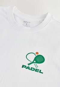 Balta medvilninė marškinėliai su laisvu siluetu, su žalia "PADEL" grafika, raketės ir oranžinio kamuoliuko detale priekyje.