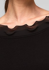 Närbild av en kvinnas nacke och nyckelben som bär en svart topp med en genomskinlig, volangkrage.