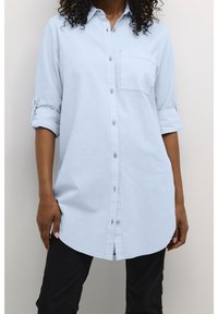 Chemise bleu clair en tissu doux, avec une poche poitrine, des manches retroussables et un ourlet incurvé. Portée avec un pantalon noir.