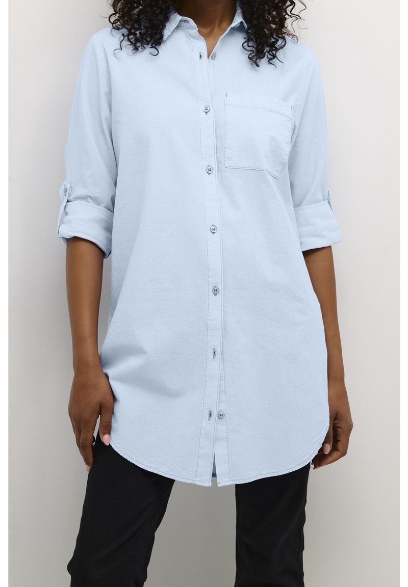Chemise bleu clair en tissu doux, avec une poche poitrine, des manches retroussables et un ourlet incurvé. Portée avec un pantalon noir.