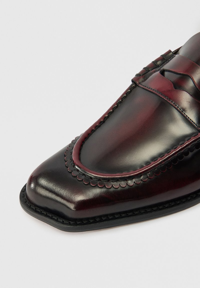 Mocassino in pelle bordeaux scuro lucido da uomo con punta quadrata, bordi decorativi a scallop e una classica fascia penny su sfondo bianco.