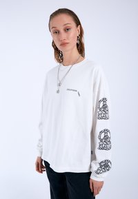 White long-sleeve t-shirt made of cotton. Features black graphic text on the front and floral designs on both sleeves. Relaxed fit.