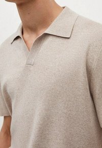 Polo en maille beige avec un col plat, des manches courtes et une texture subtile. Présente une patte de boutonnage discrète au niveau de la poitrine.