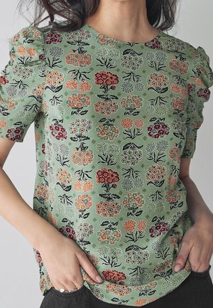 Zielona, kwiecista bluzka z krótkim rękawem i bufiastymi ramionami noszona przez osobę z rękami w kieszeniach czarnych spodni.