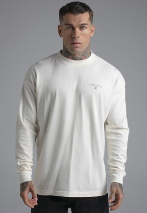 Muž s tetovaniami na krku a ruke nosí biele tričko s dlhým rukávom so logom "SIKSILK" a stojí pred jednoduchým sivým pozadím.